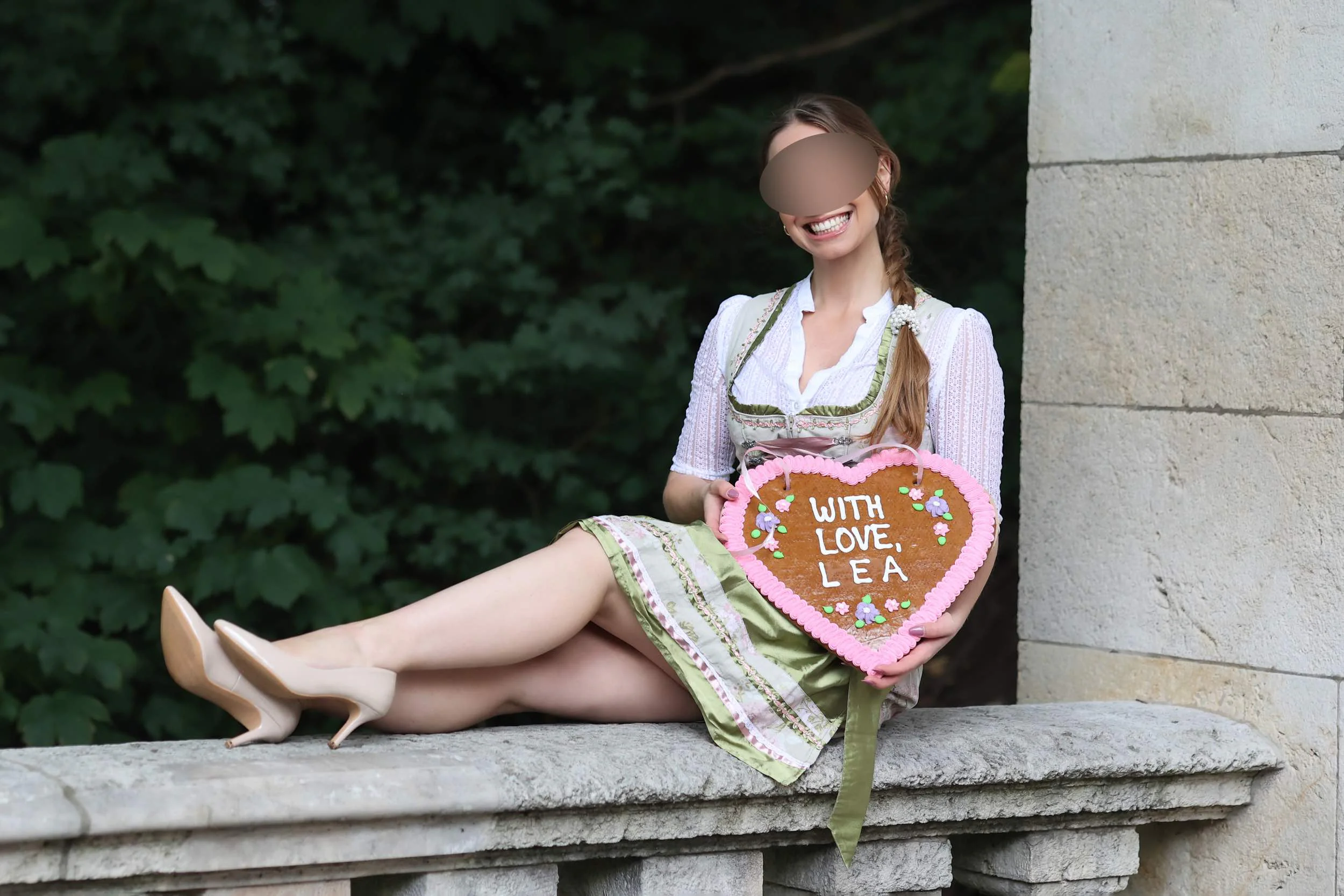 Escort Juna im Dirndl mit Lebkuchenherz auf der Balustrade in München