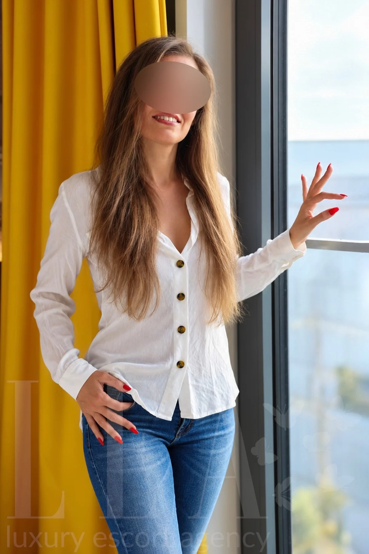 Escort Dame Leto in Heidelberg in weißer Bluse und Jeans am Fenster